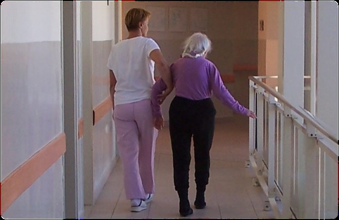 Starija osoba hoda niz hodnik doma za starije, pridržava je zaposlenica doma